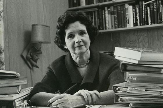Black and white photo of Rachel Carson sitting behind a desk full of books with pen in hand. 